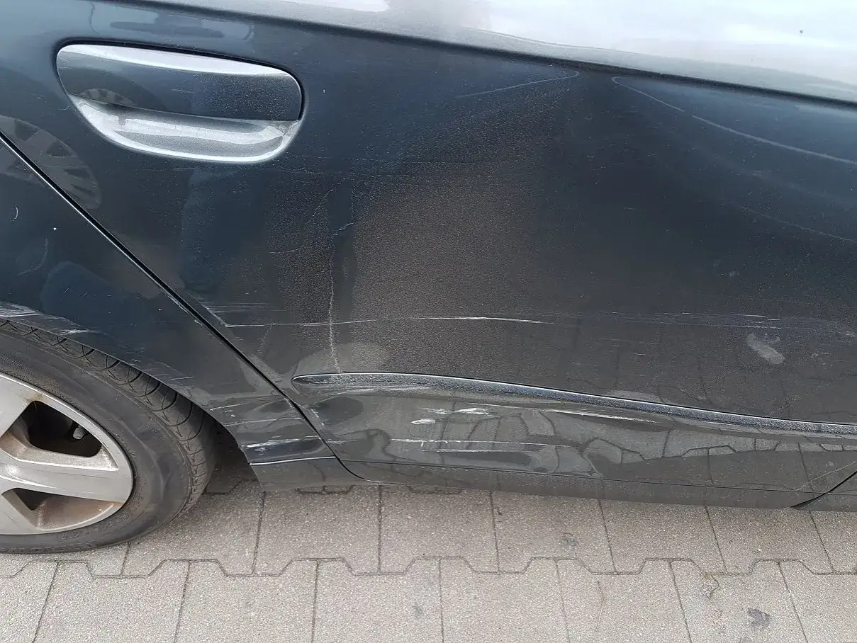 Zarysowany błotnik w aucie, widoczne liczne zadrapania i wgniecenia na ciemnym lakierze.